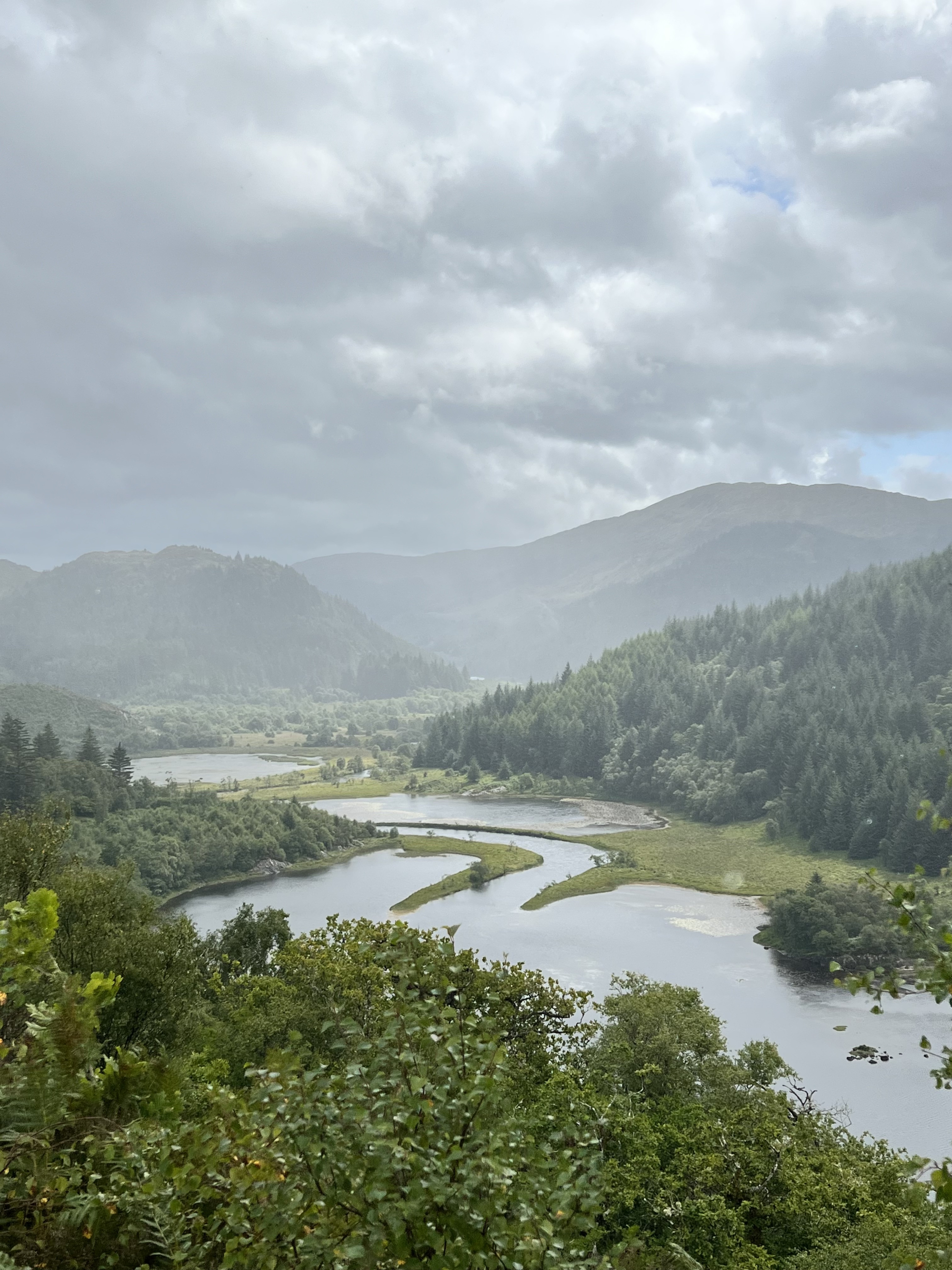 A river winds through a misty wooded valley seen from above, hills rising on either side under a cloudy sky.