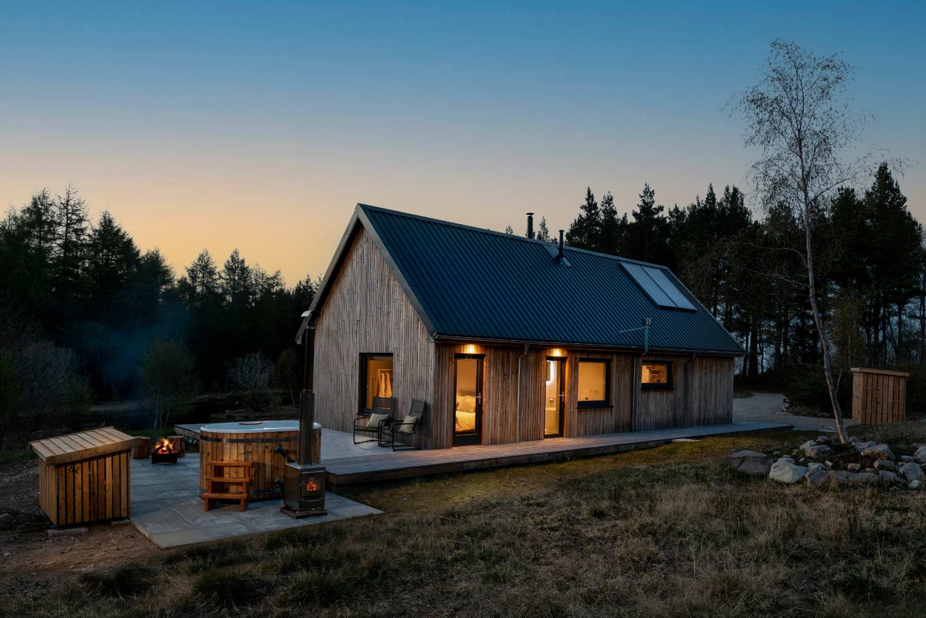 A timber cabin glows with lit windows at dusk, smoke rising from a wood-fired hot tub on the deck beside a fire pit.