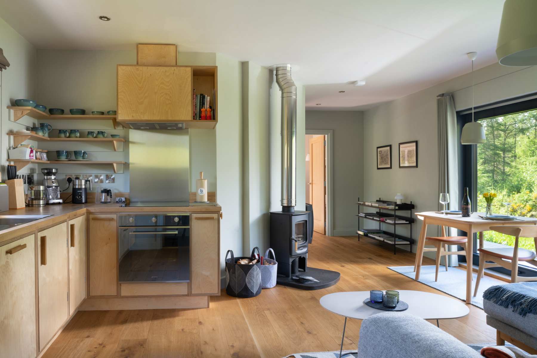 An open-plan kitchen and living area with a wood burning stove and flue, shelves of crockery and a dining table by the window.