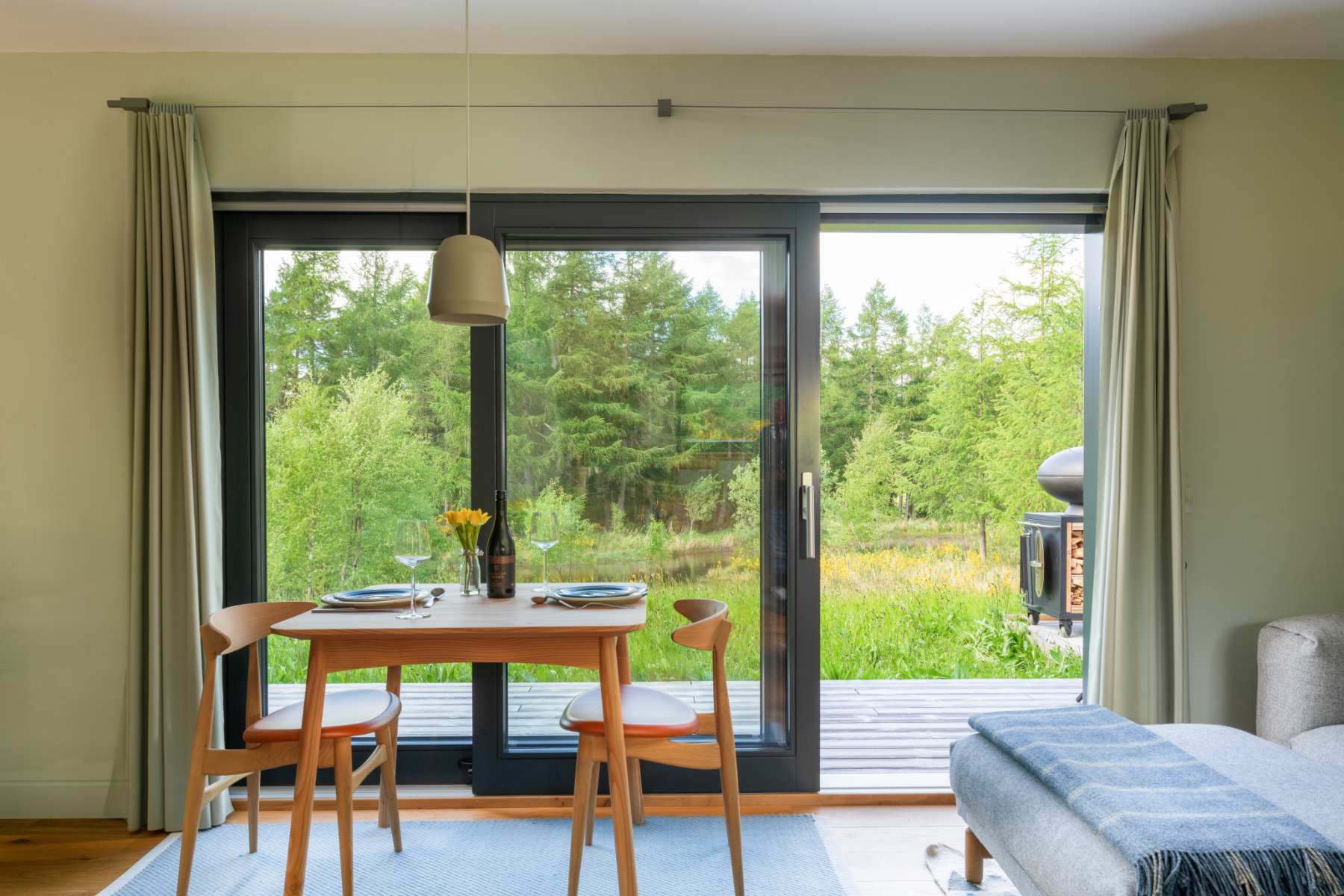 A round table set for two stands before open glazed doors looking onto summer woodland and a deck.