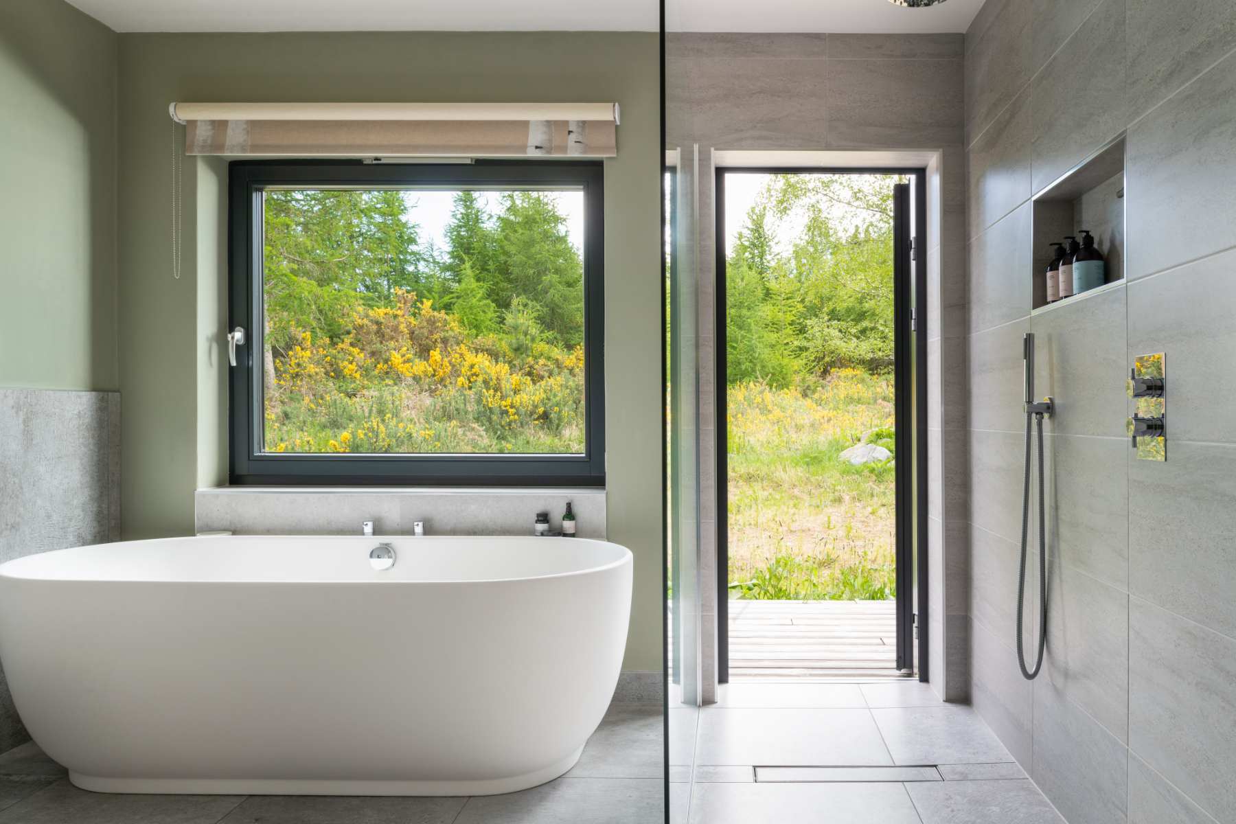 A freestanding oval bath stands before a window onto flowering gorse, a walk-in shower and open door to the garden alongside.
