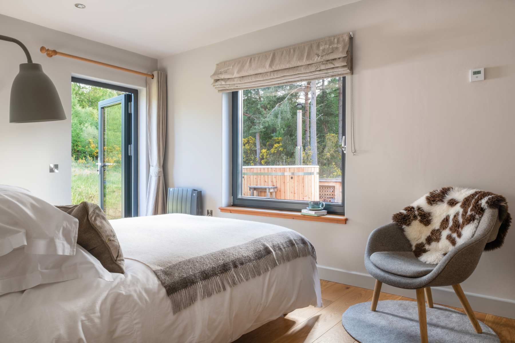 A bed with white linen faces glazed doors open onto a deck, a sheepskin draped over a grey armchair to the side.