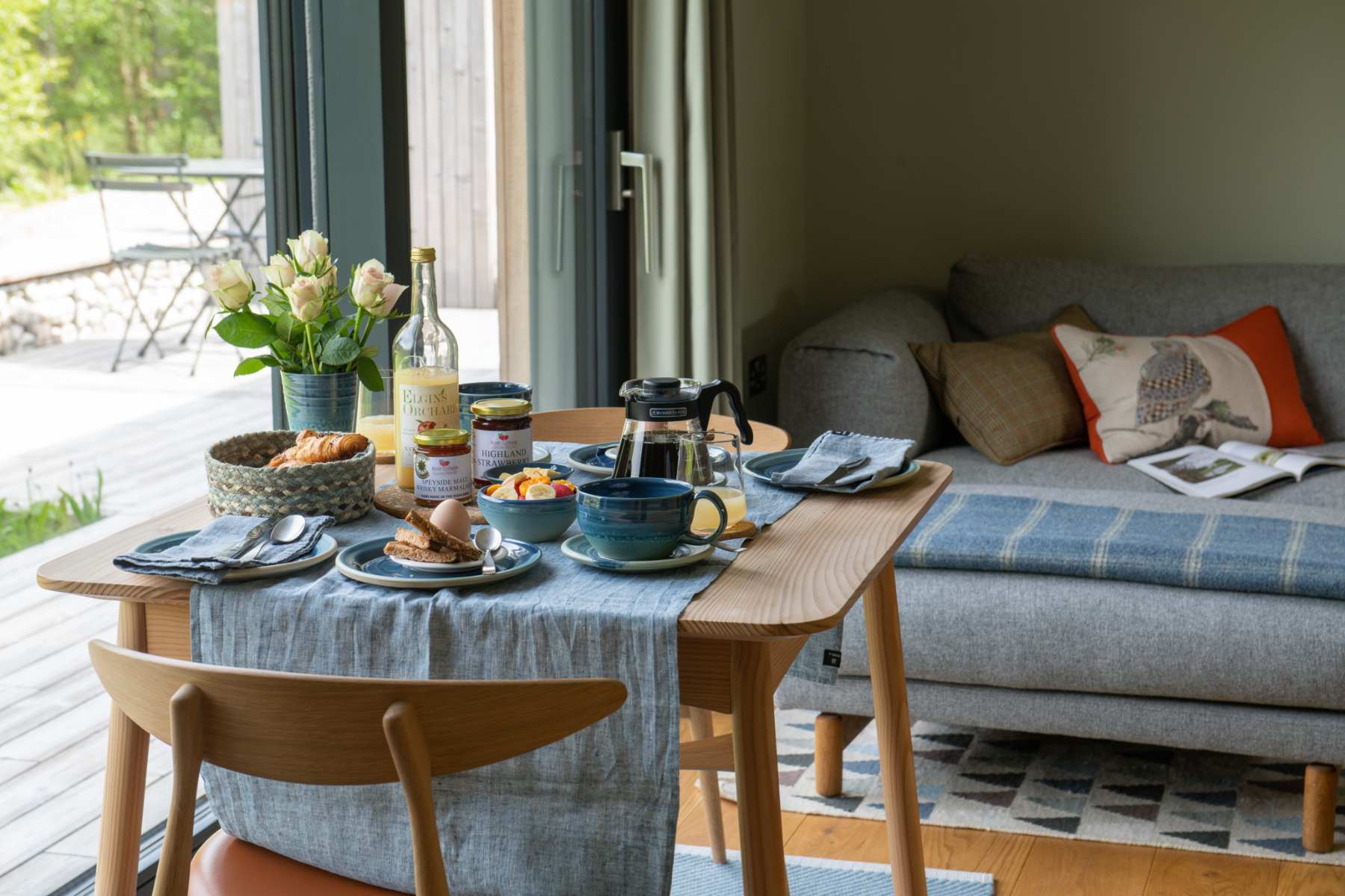 A table laid with breakfast items and flowers stands before a grey daybed with patterned cushions, glazed doors open onto a deck.