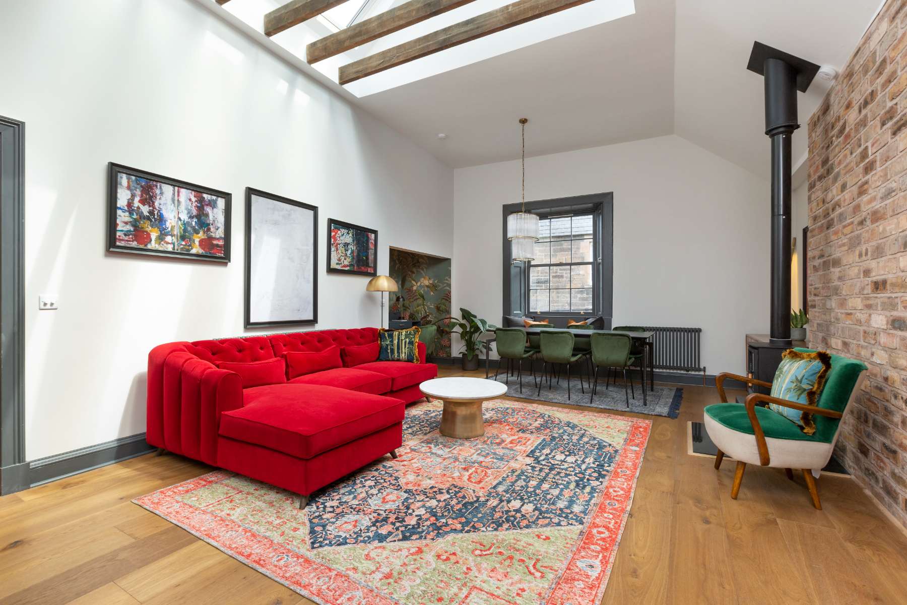 A red corner sofa sits before a wood-burning stove and exposed brick wall, a gallery of framed prints on the opposite wall and a dining table beyond under a skylight.