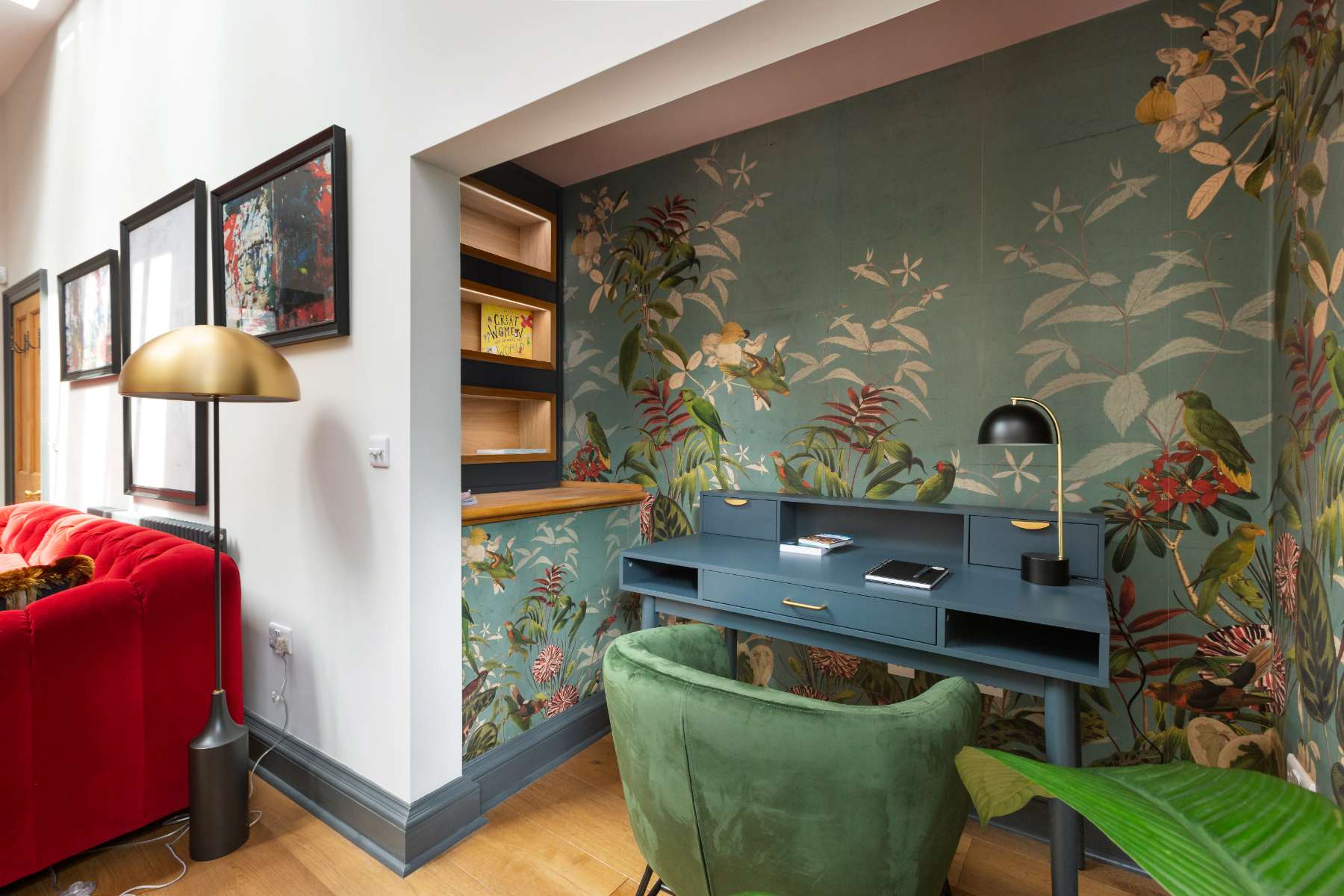 A dark blue desk with a lamp and books sits before botanical mural wallpaper, a green velvet chair and gold floor lamp completing the nook.