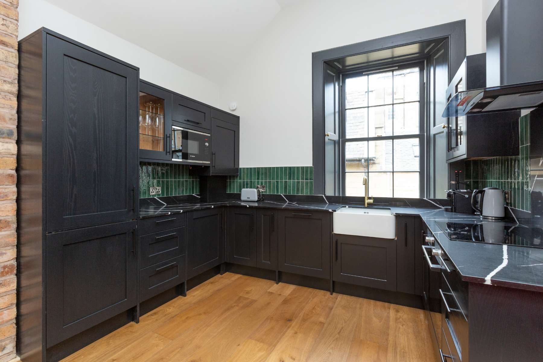 A black kitchen with a green tiled splashback has a Belfast sink with a gold tap beneath an arched window.