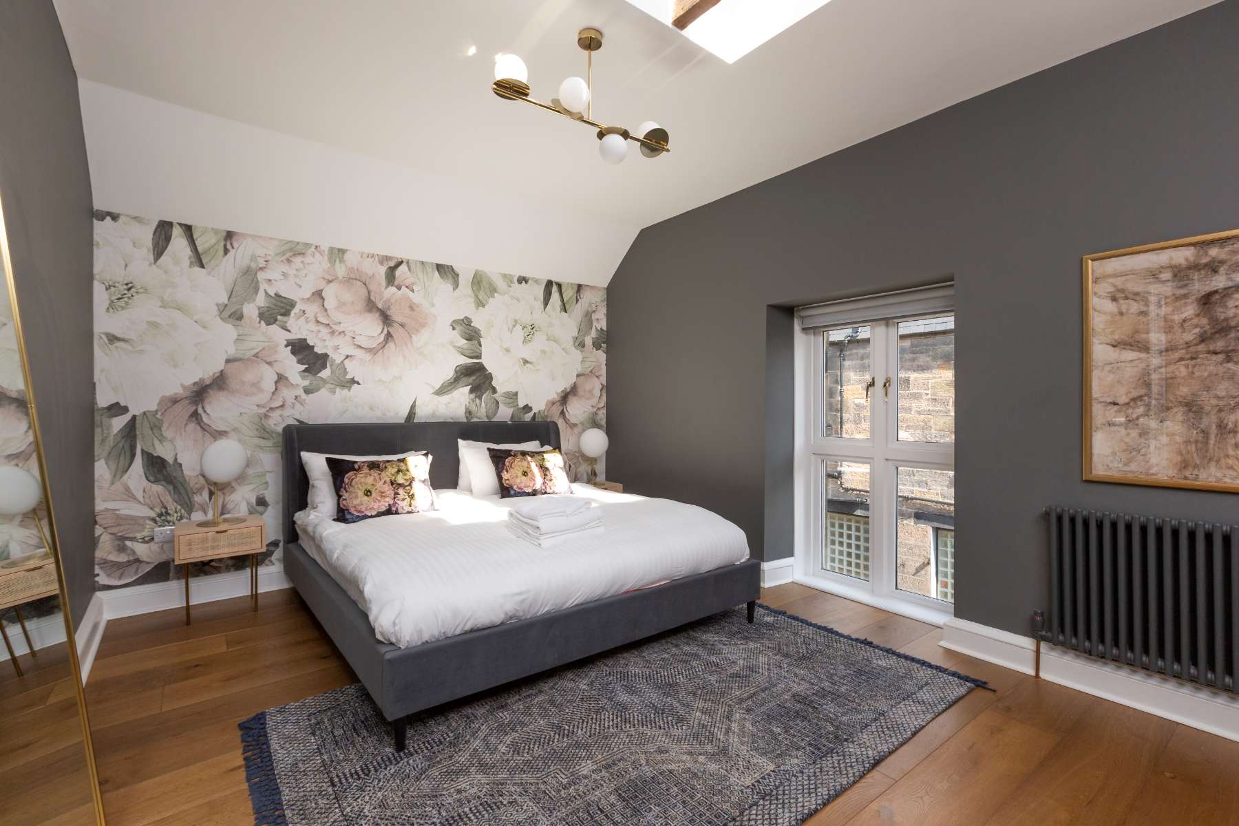 A grey upholstered bed with patterned cushions sits against a floral mural wall, a glass door leading to a balcony beside a radiator.