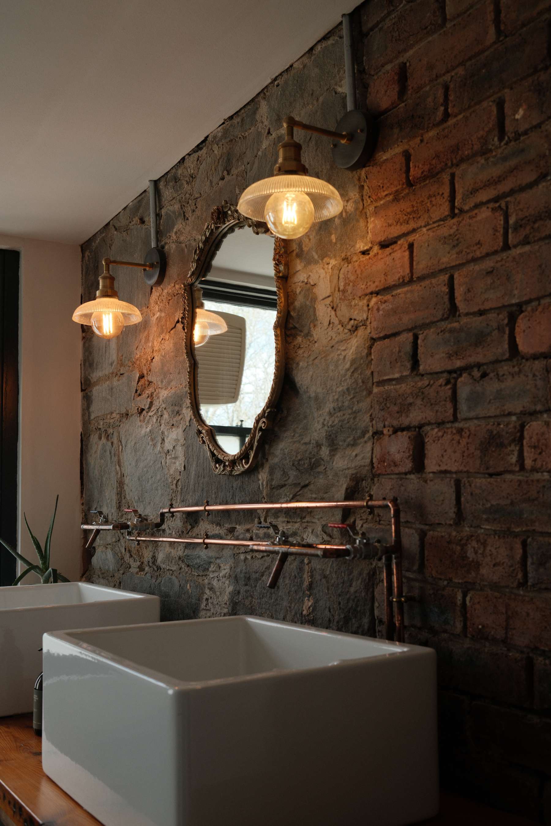 Twin basins stand below an ornate gilt mirror flanked by wall lights, set against an exposed brick and stone wall with copper pipework.
