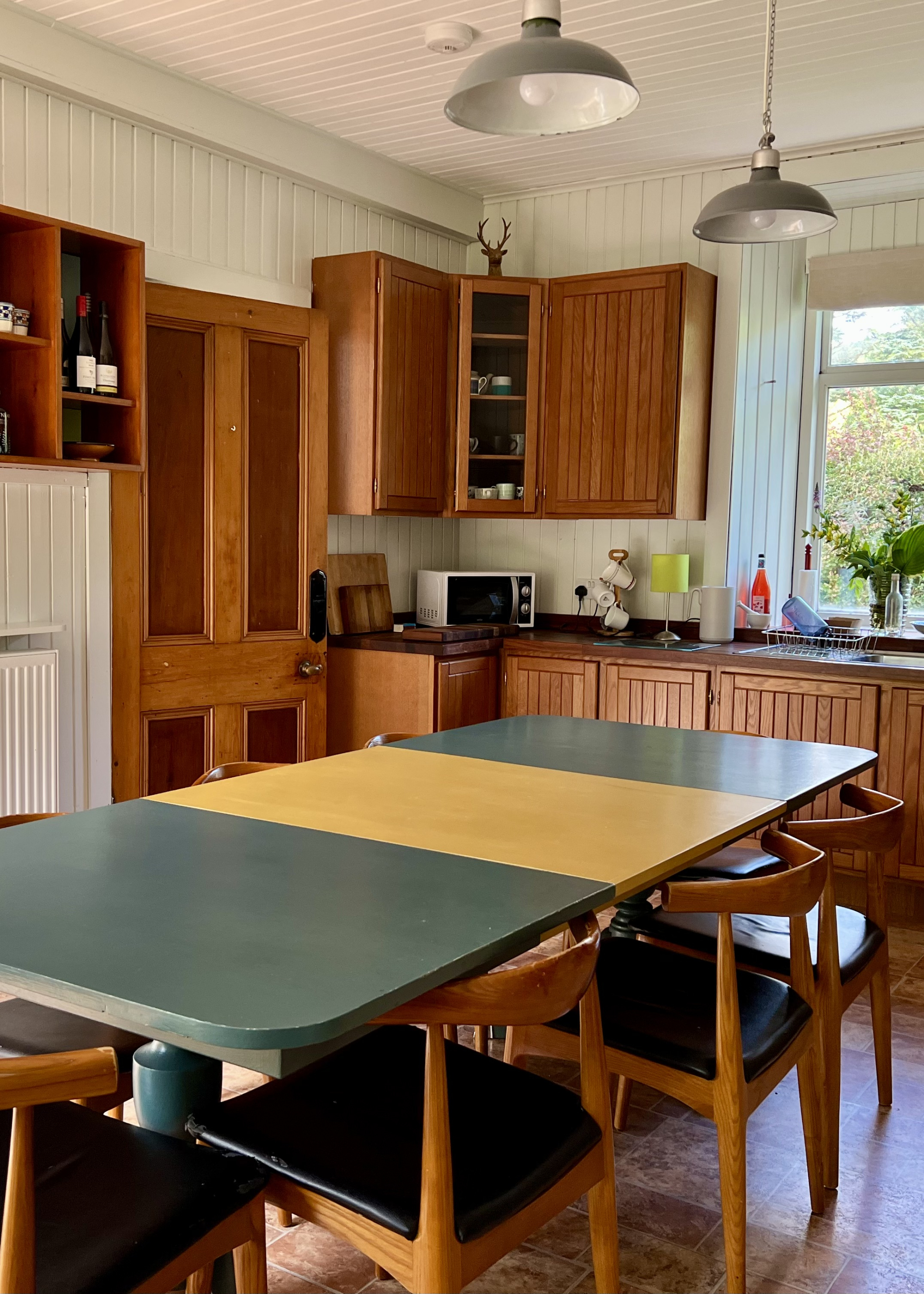 A kitchen with wooden cabinetry and a green and yellow dining table, a microwave and glass-fronted cupboard display crockery.