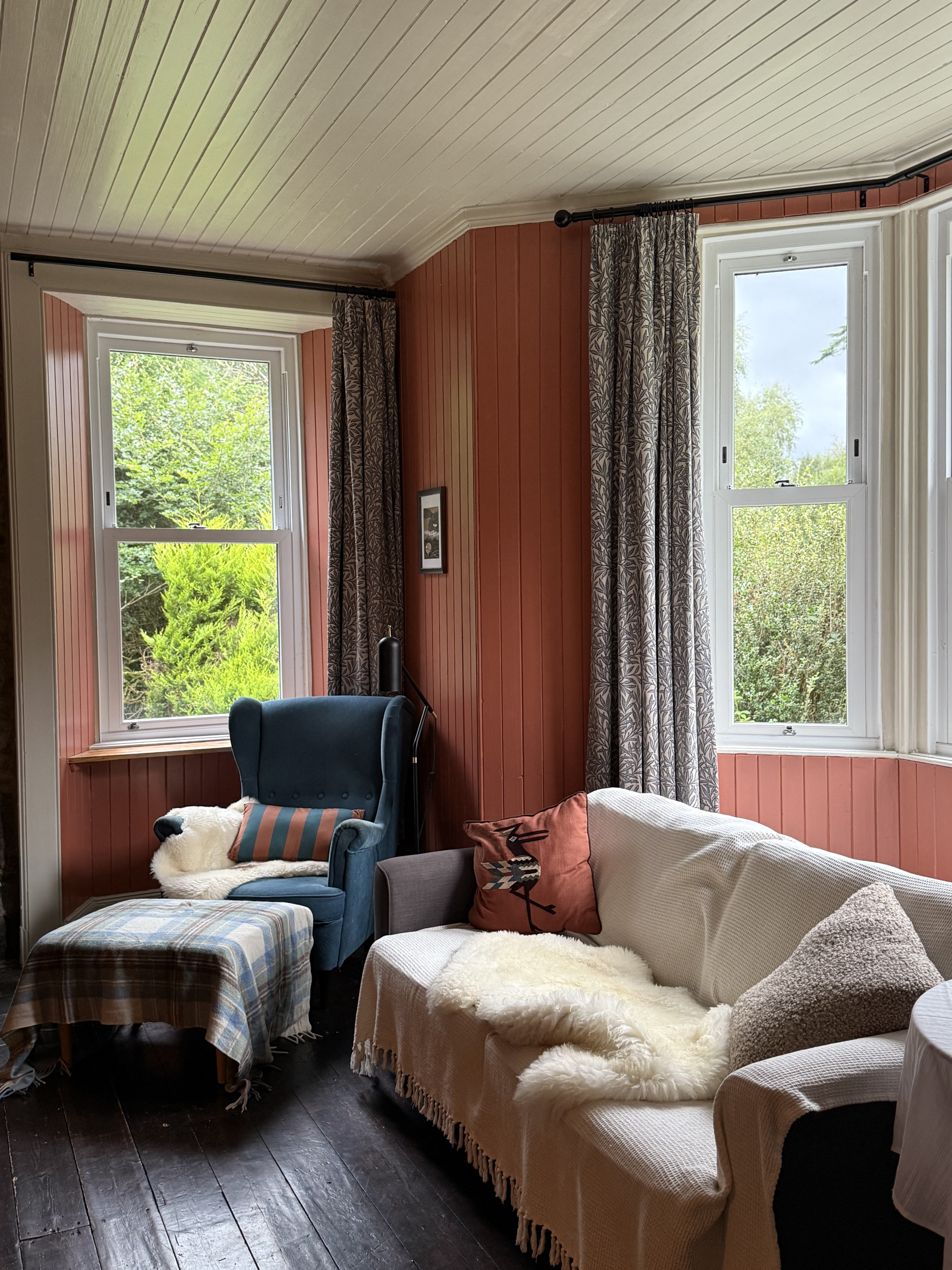 A terracotta-panelled sitting room with a blue wing-back armchair and a sofa draped in sheepskin throws, floral curtains framing two windows.