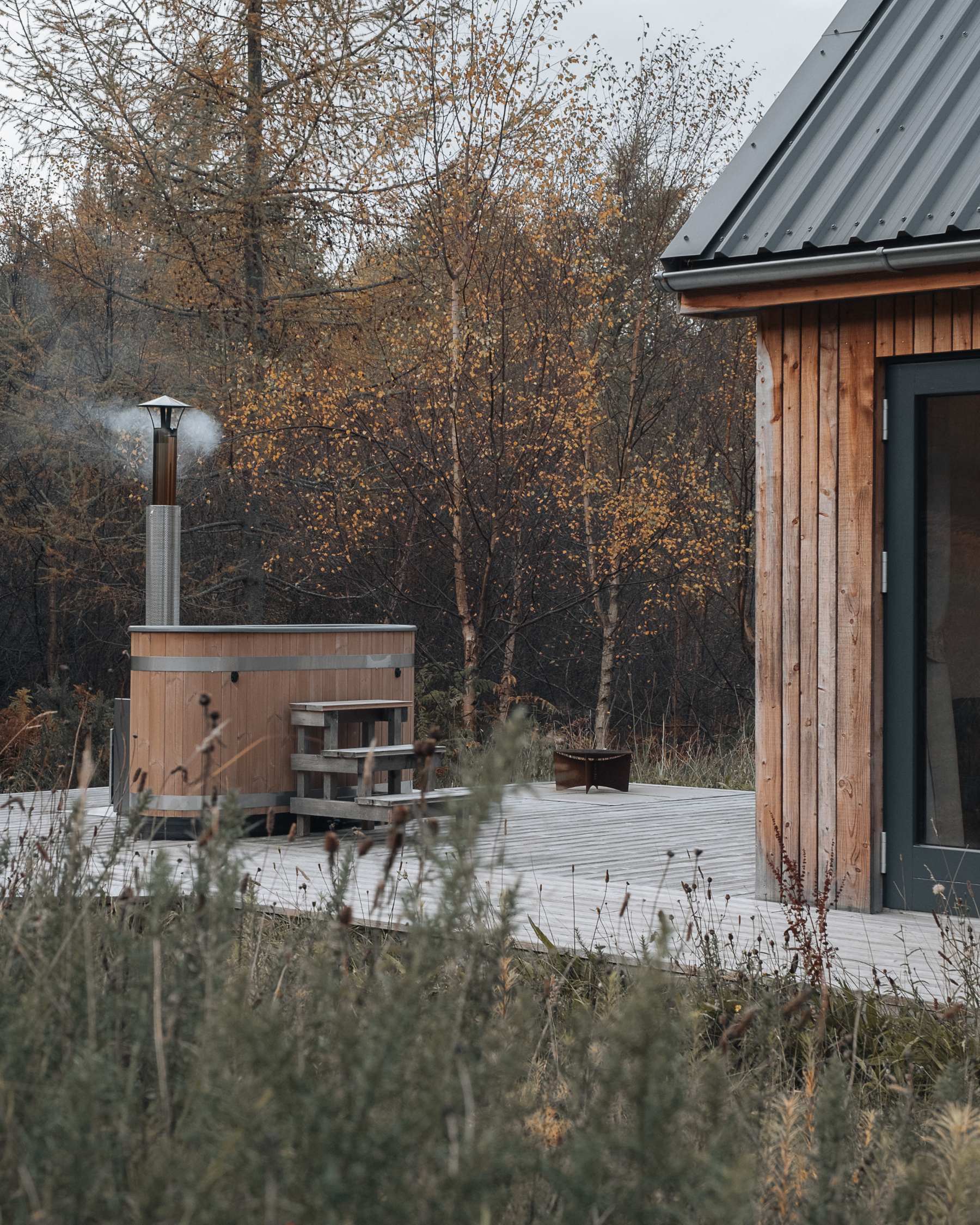 A wood-fired hot tub steams on a deck beside the timber cabin, autumn birches turning gold behind.