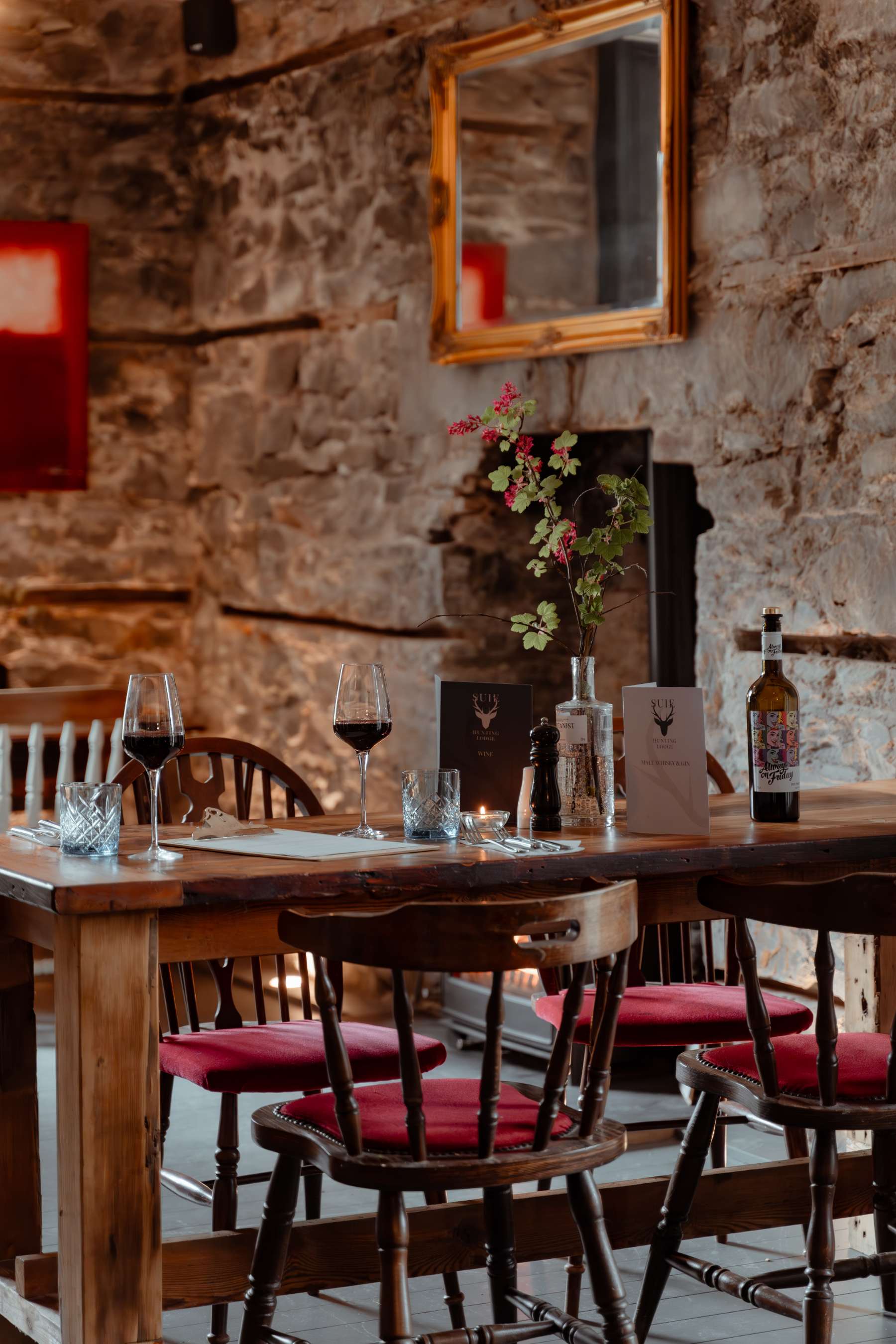 A long wooden table set with red wine and menus stands before a stone wall hung with a gilt mirror, red-cushioned chairs arranged around it.