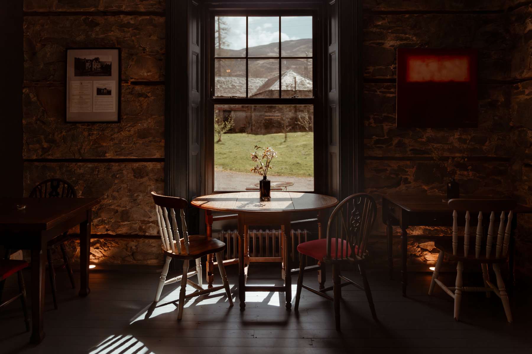 A round table set with a wine bottle and a small vase of flowers stands before a window framing hills, wooden chairs with red cushions around it.