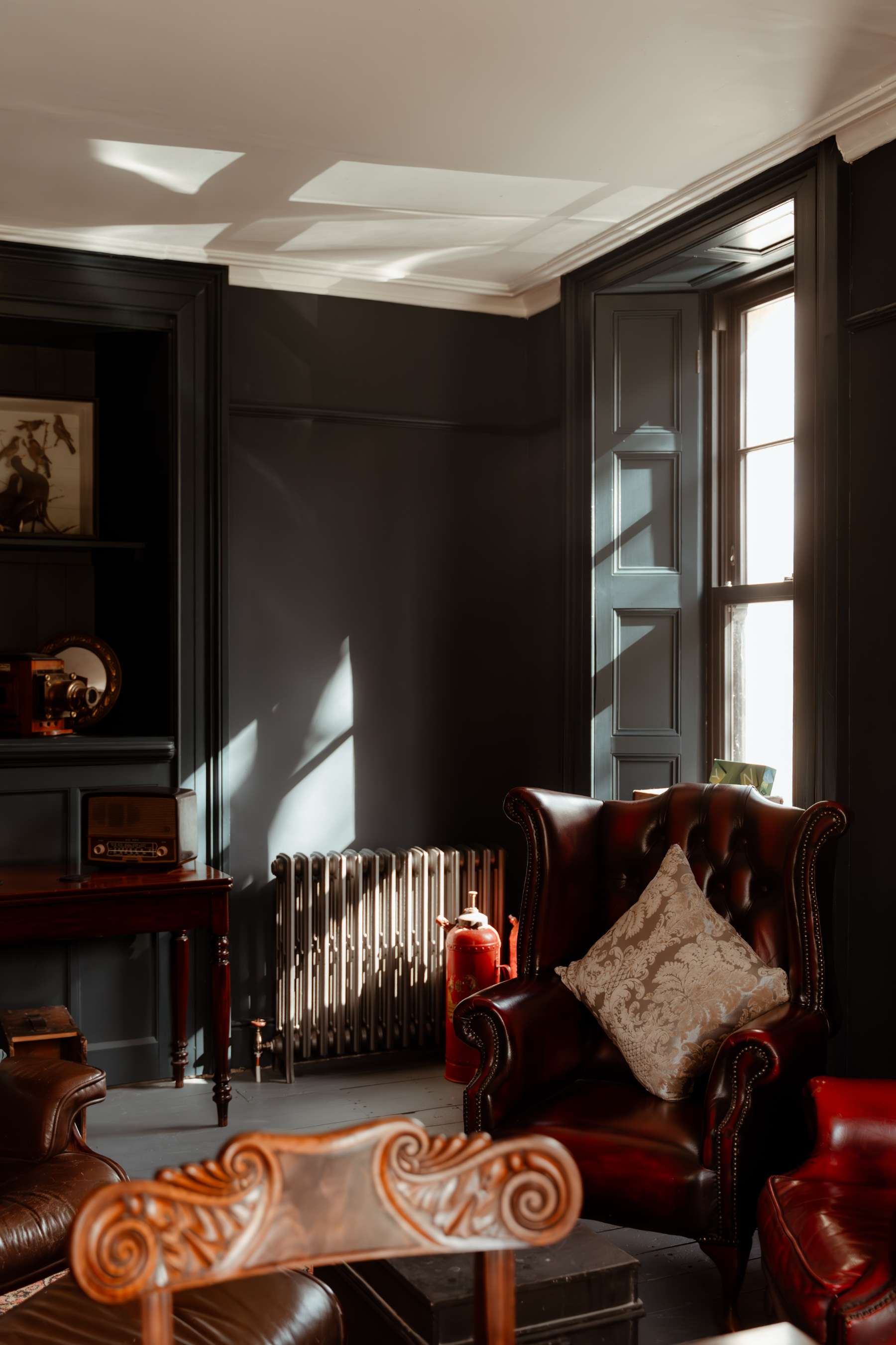 Leather wing-back armchairs sit before a curtained window in a dark green panelled room, a radiator and vintage radio on a side table nearby.