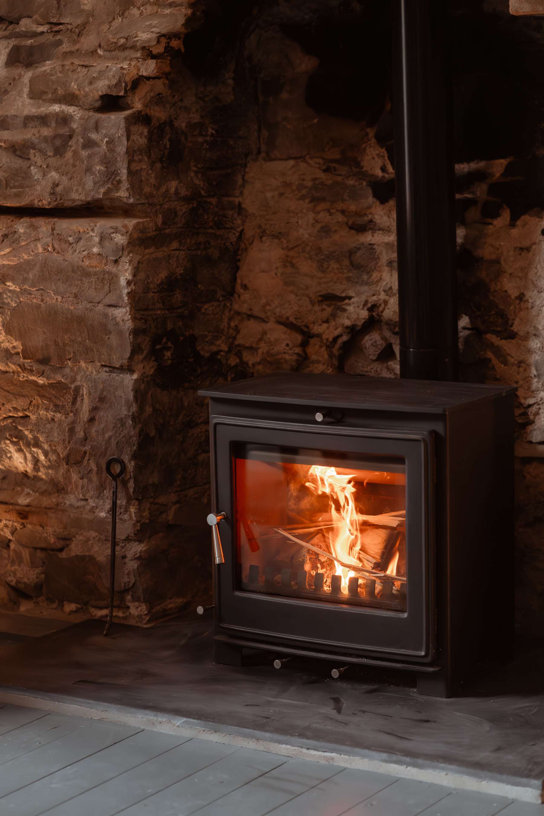 A wood burning stove burns brightly beneath a black flue, set into a rough stone alcove with a companion set beside it.