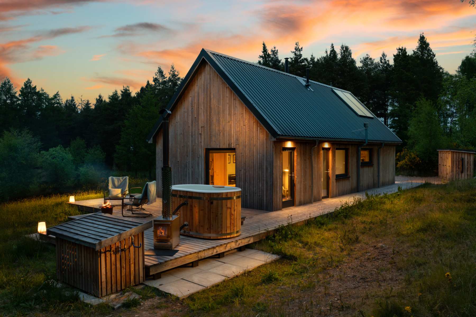 The timber cabin lit at dusk against a pink sky, a hot tub steaming on the deck beside two chairs.