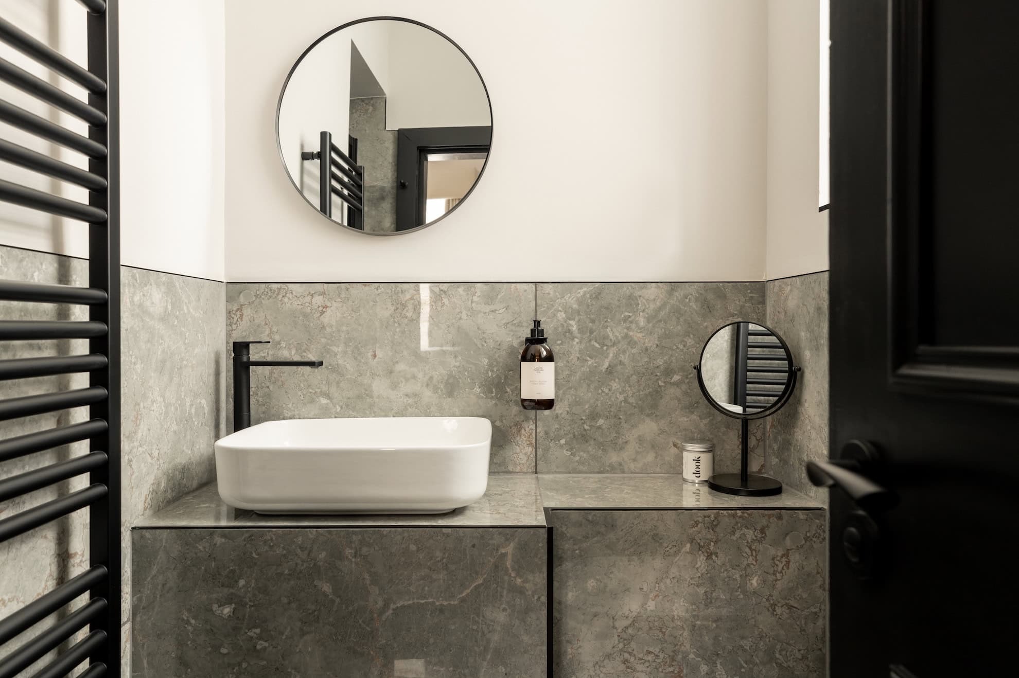 A round mirror hangs above a rectangular basin set into a grey marble vanity, a pump bottle and small round mirror beside it.