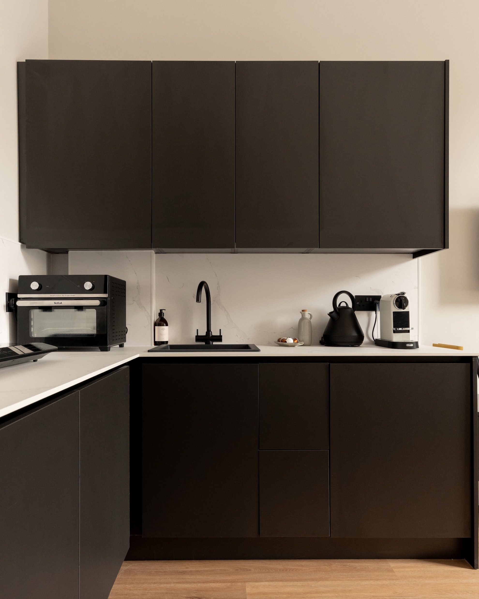 A matte black kitchen with an integrated oven and hob, a kettle and coffee machine on the counter beside a black tap.