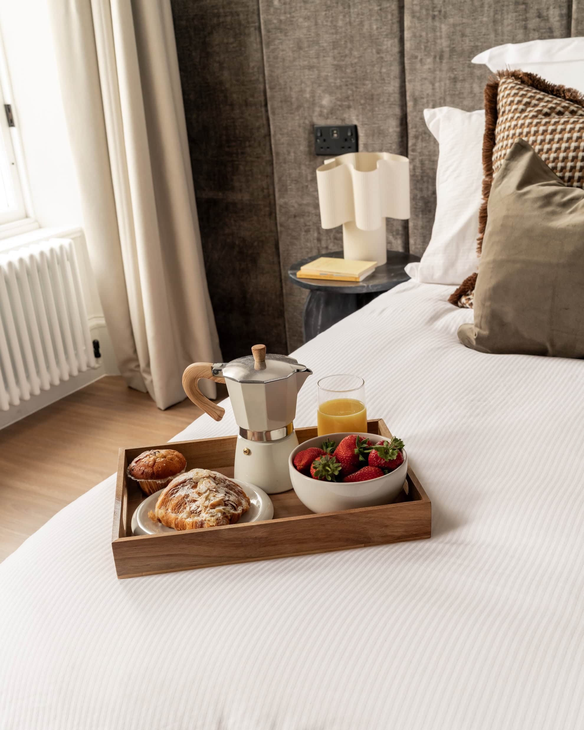 A stovetop coffee pot, pastries and a bowl of strawberries sit on a wooden breakfast tray placed on a bed.