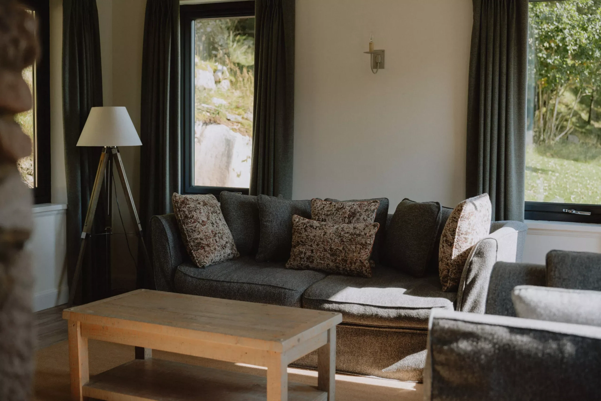 A grey corner sofa with floral cushions sits before curtained windows looking onto greenery, a floor lamp and wooden coffee table completing the room.