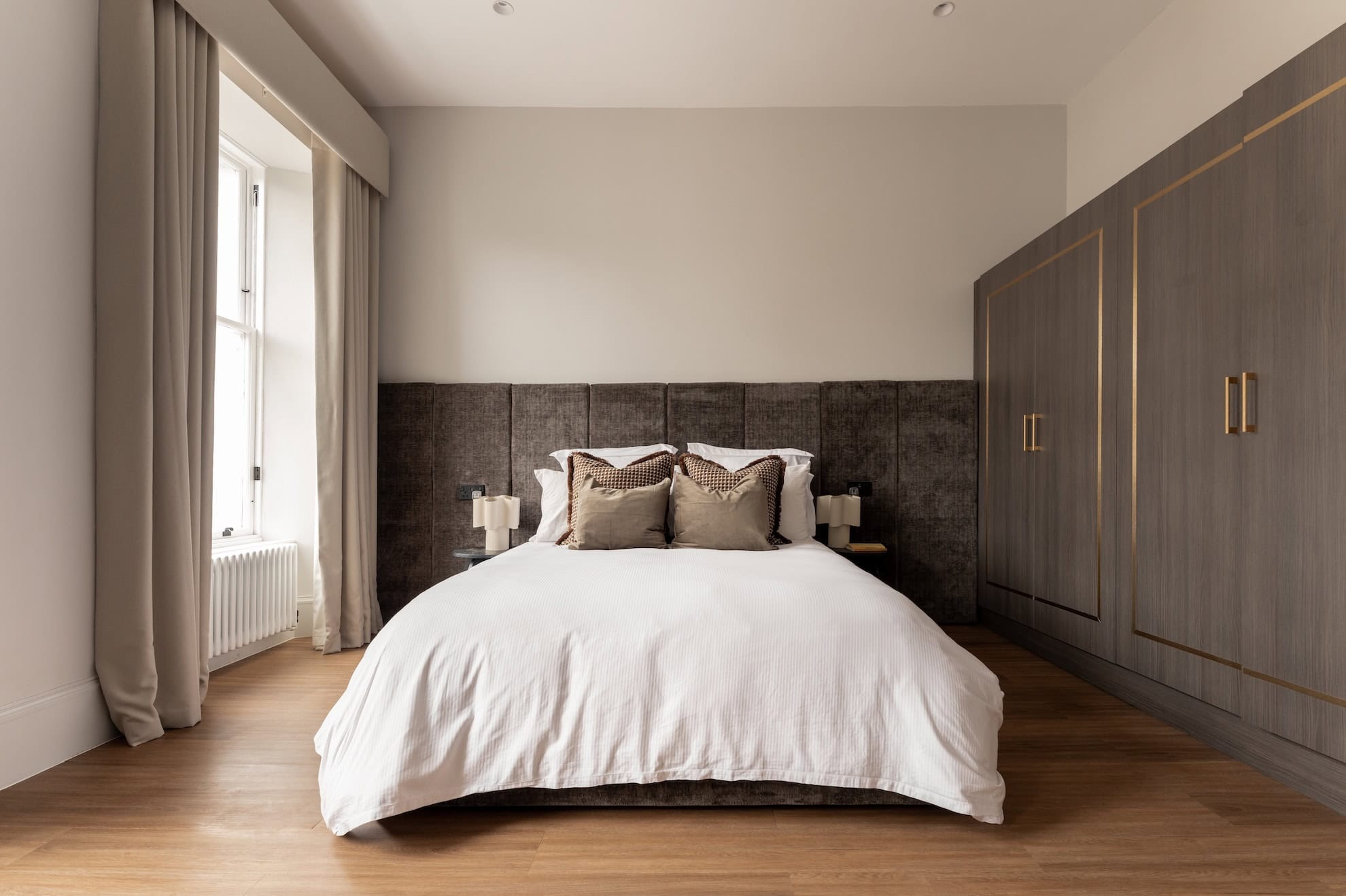 A bed with a dark upholstered headboard and matching cushions sits before a curtained window, fitted wardrobes with brass handles to the right.