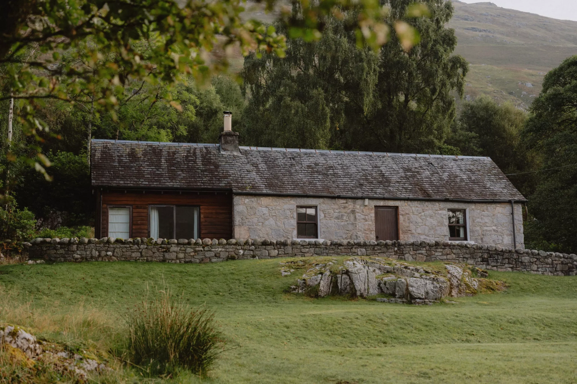 A stone cottage with a dark timber-clad extension stands behind a drystone wall and lawn, trees and hills rising behind.