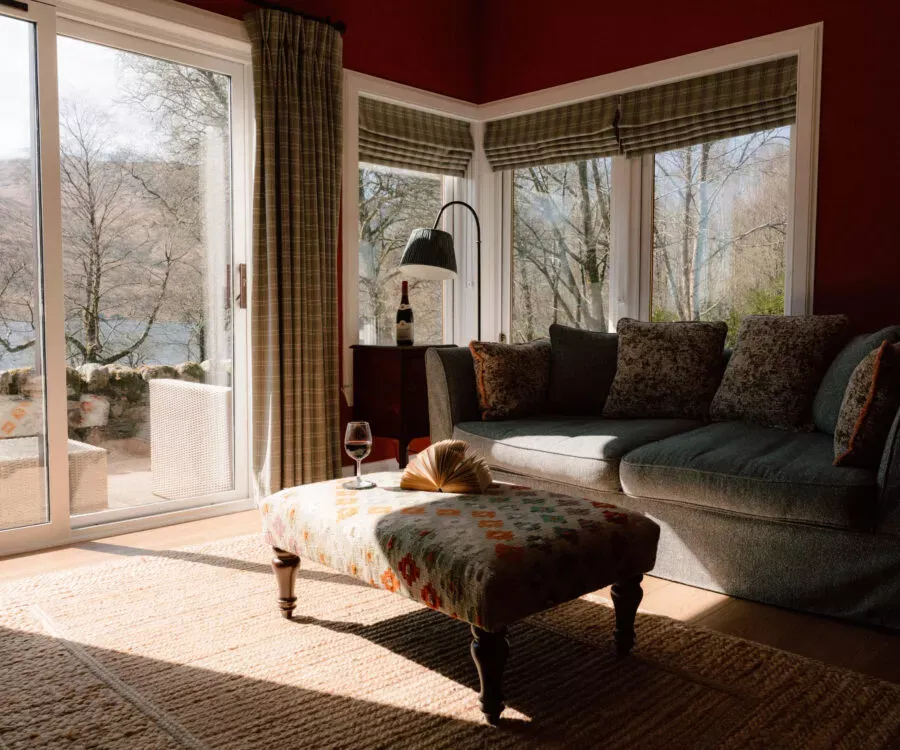 A patterned ottoman sits before a green sofa with floral cushions, a gooseneck lamp beside a glass door looking out to the loch.