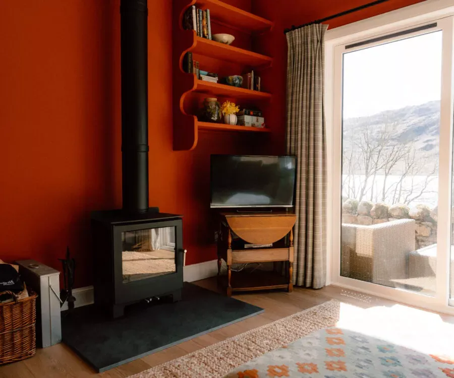 A black wood-burning stove sits in a burnt orange room beneath open shelving of books, a television on a wooden stand and a sliding glass door to the view.