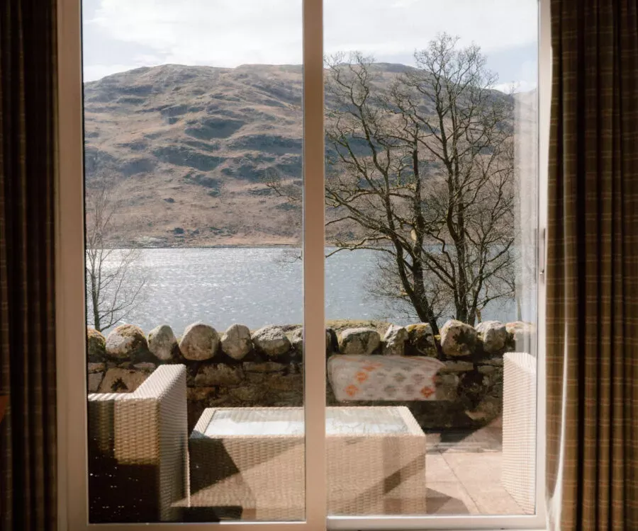 Curtains frame a glass door looking out over a loch to hills, wicker furniture and a mosaic-tiled bench set on the terrace beyond a drystone wall.