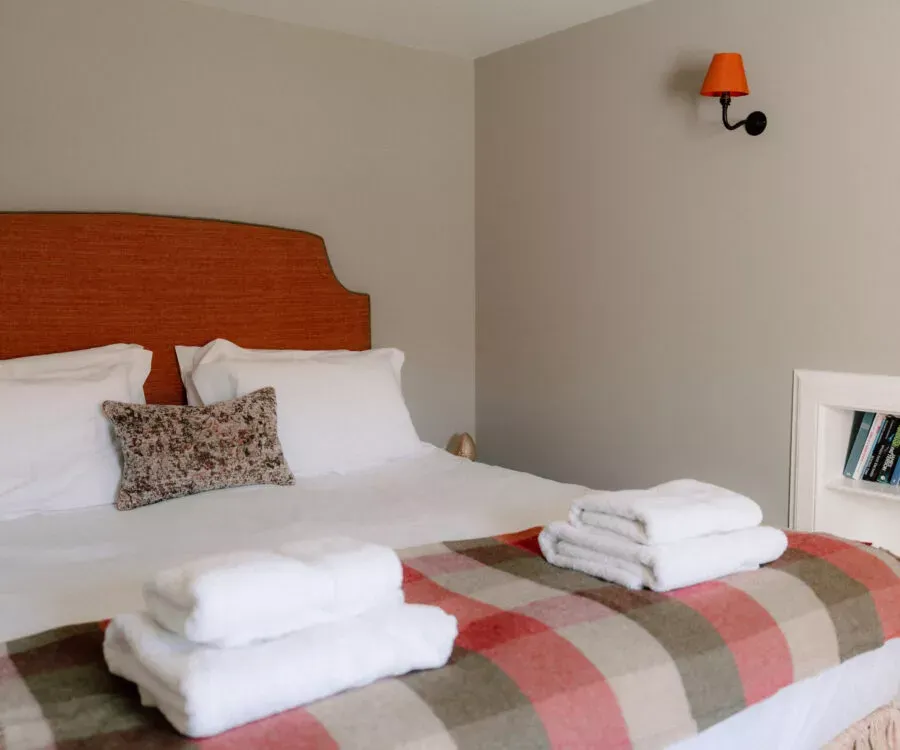 A bed with a burnt orange headboard is dressed in white bedding with folded towels and a tartan throw, a wall lamp lit beside it.
