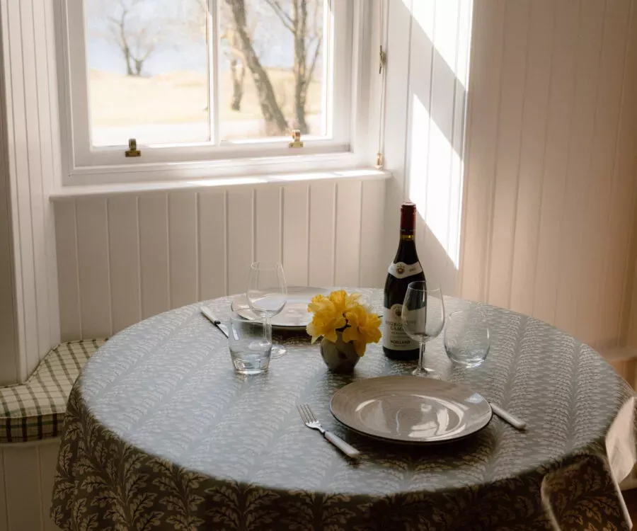 A round table set for two by a window holds a bottle of wine, glasses and a small vase of yellow flowers, a cushioned bench seat beside it.
