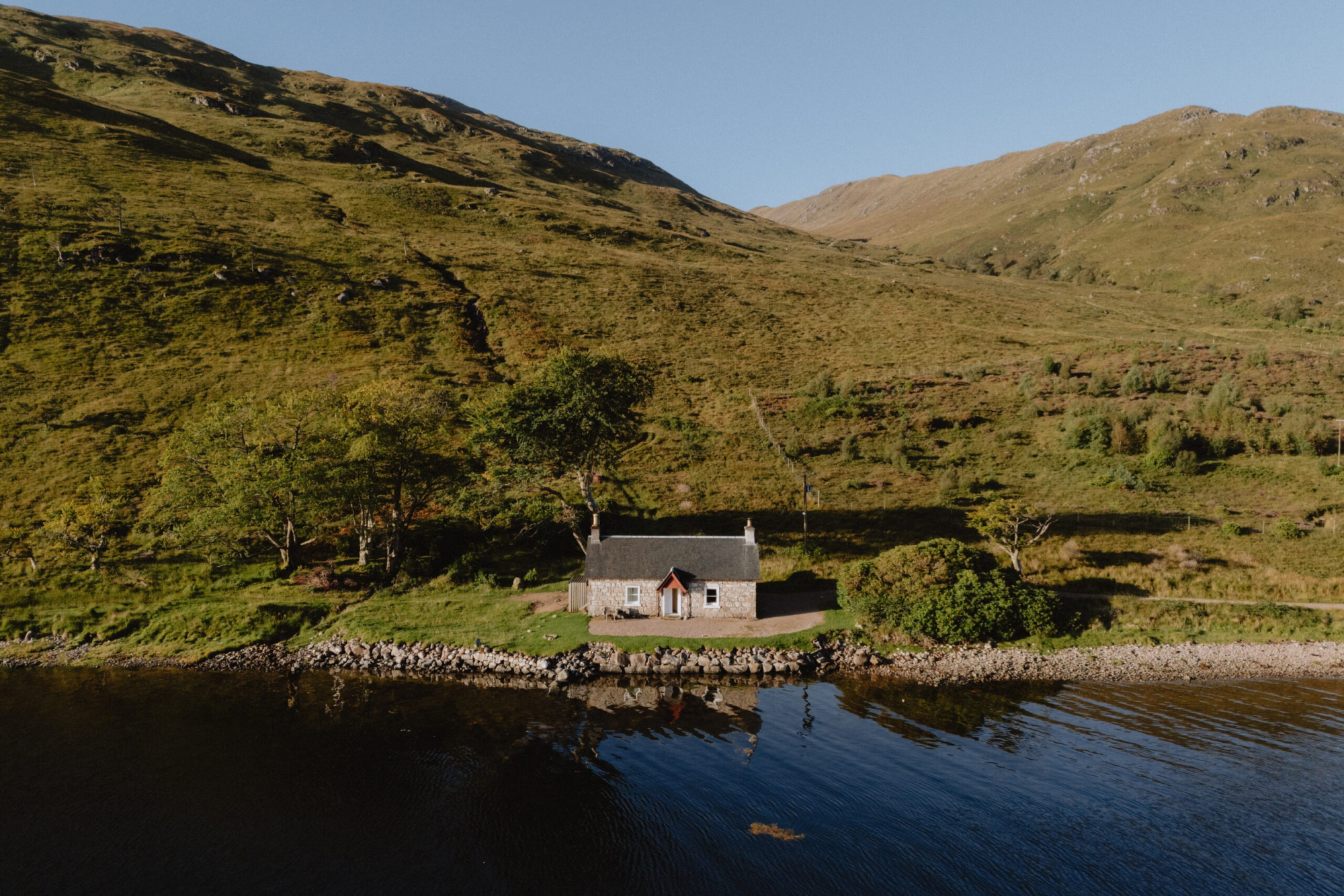 An aerial view shows a white cottage on a wooded loch shore, hills rising steeply behind it.