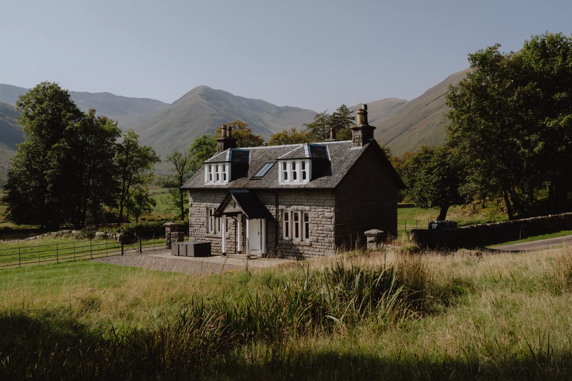 A grey stone gate lodge with dormer windows and chimneys stands amid grassy fields with hills rising behind.