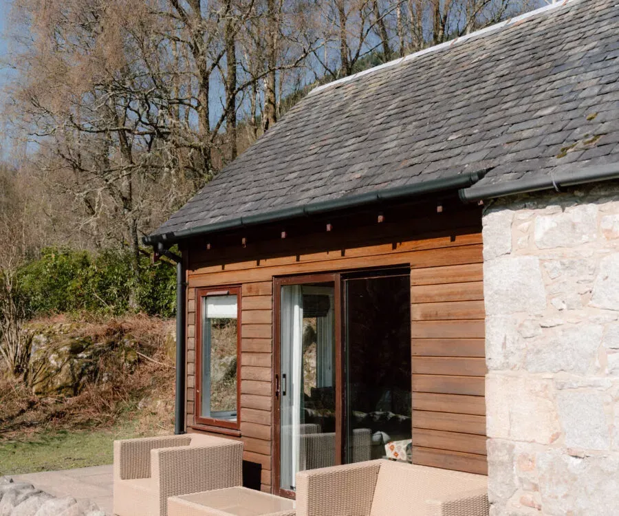 A timber-clad extension with glazed doors opens onto a paved terrace set with wicker chairs, a stone wall running alongside.