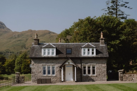 A stone lodge house with dormer windows and chimneys stands before hills, a gravel driveway leading to the lawn.