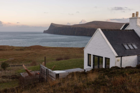 A white house with a dark slated roof sits above a rocky headland and sea, a weathered bench in the garden.