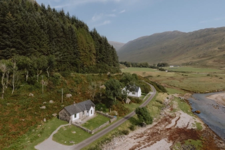 An aerial view shows a white cottage and stone bothy beside a road running along a wooded glen and river.
