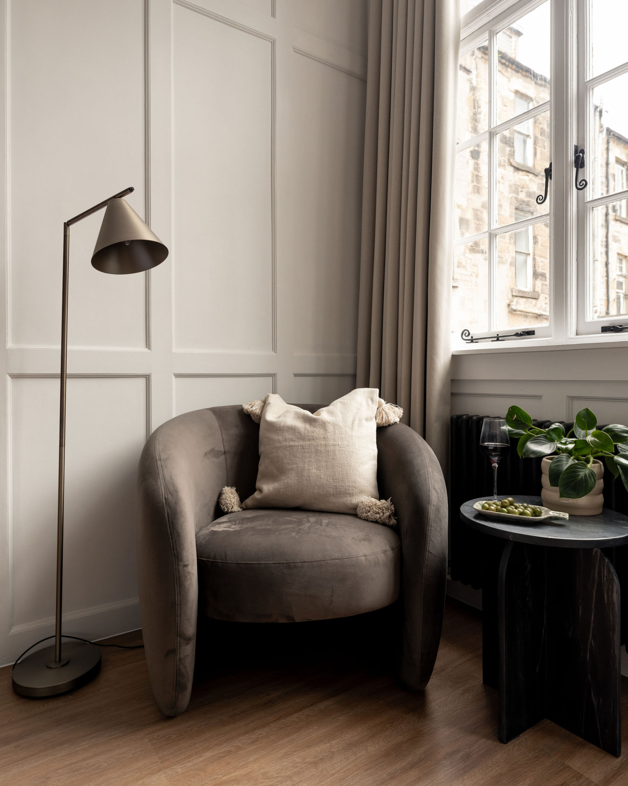 A grey velvet armchair with a cushion sits beside a floor lamp and round black side table, a window looking onto stone buildings.