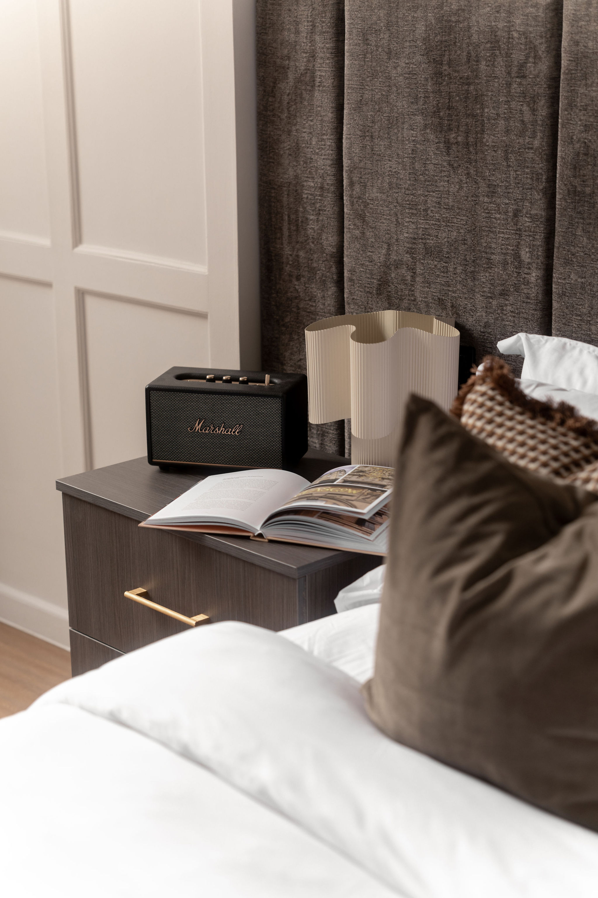 A black speaker and a ridged lamp stand on a bedside table beside an open book, a brown cushion resting on the bed.