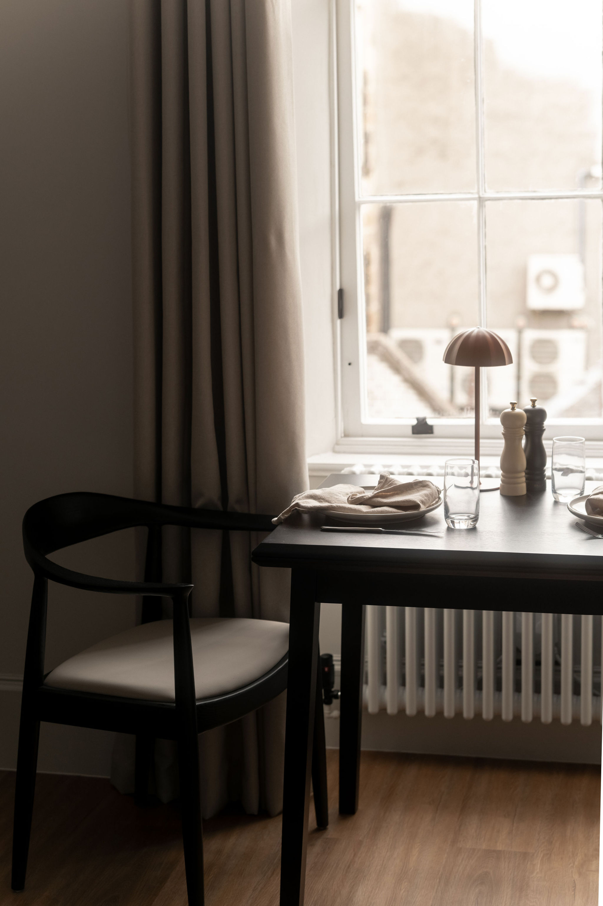 A black dining chair stands beside a table set with a lamp, pepper grinder and glasses before a tall window.