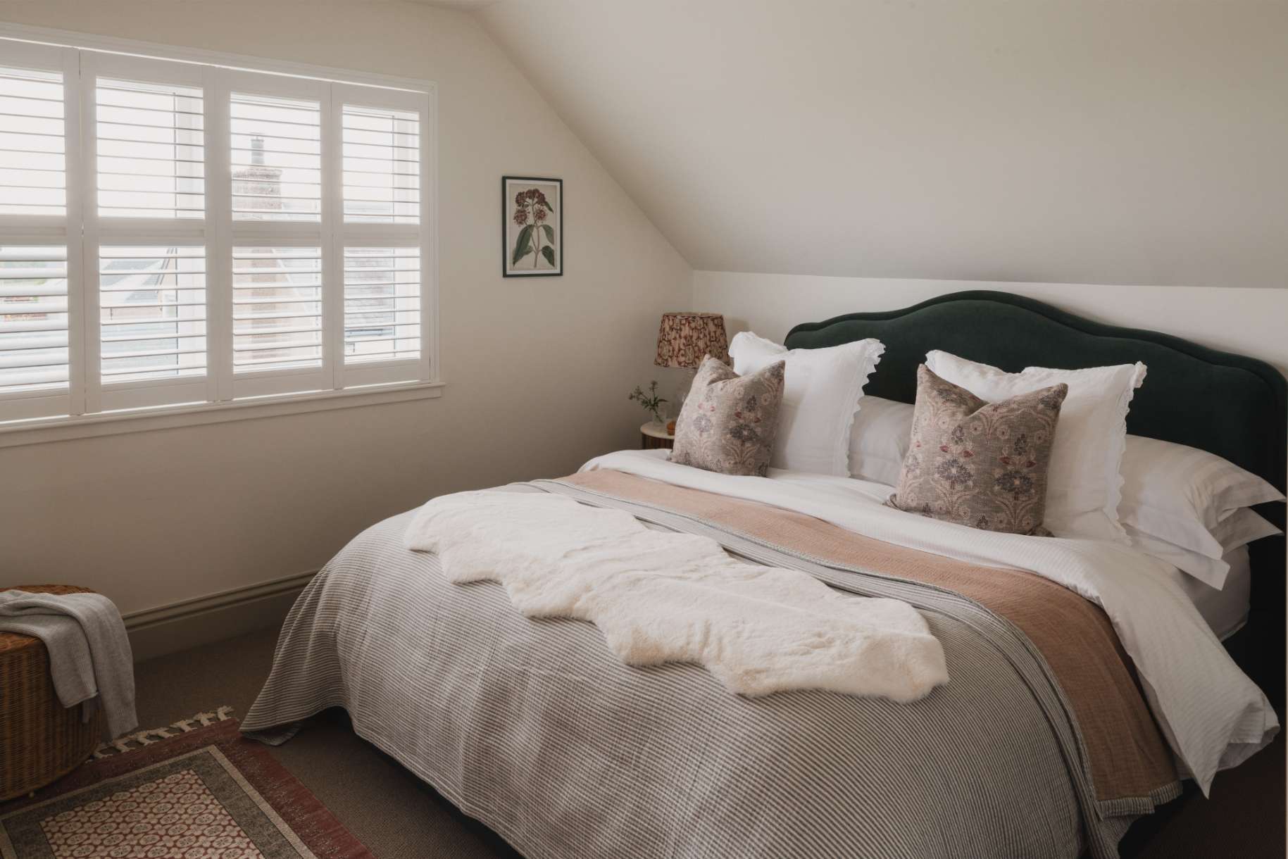 A bed with a dark green headboard and patterned cushions sits below a shuttered window, a sheepskin throw at the foot.
