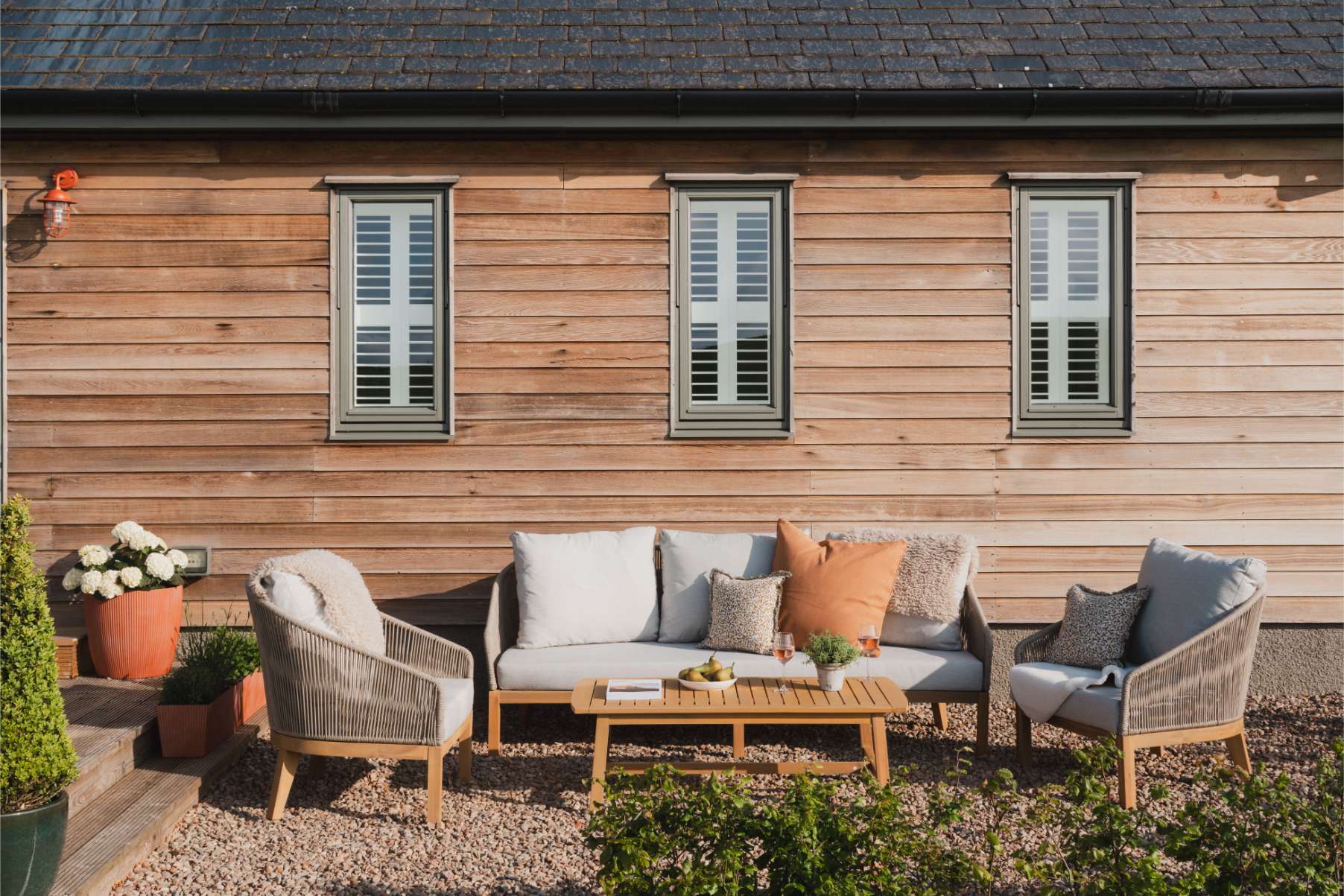 A wicker sofa with orange cushions sits before a green kitchen with a butler's sink, patterned armchairs and a low table in the foreground.
