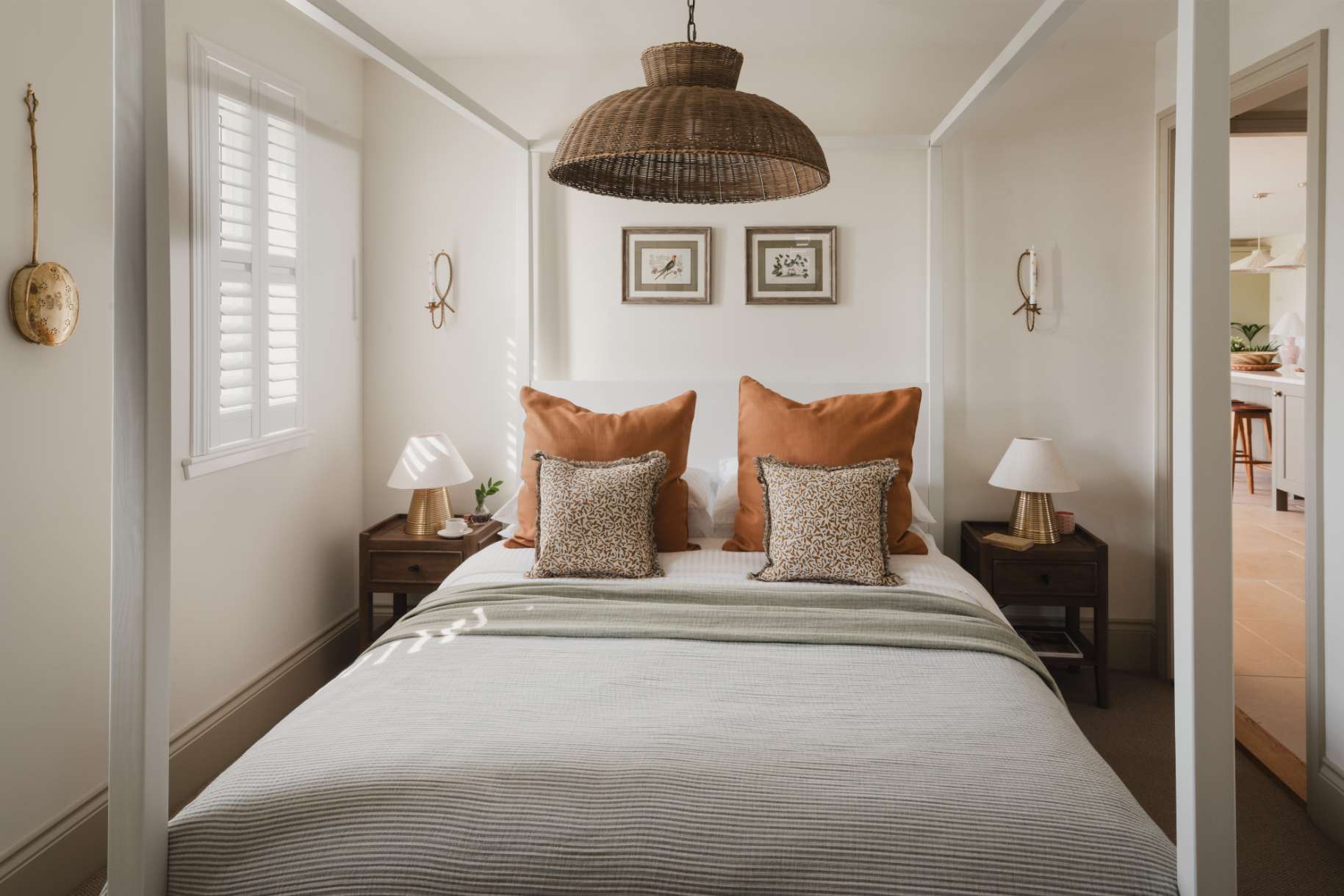 A four-poster bed with orange cushions sits beneath a woven pendant light, framed prints on the wall and matching lamps on the bedside tables.