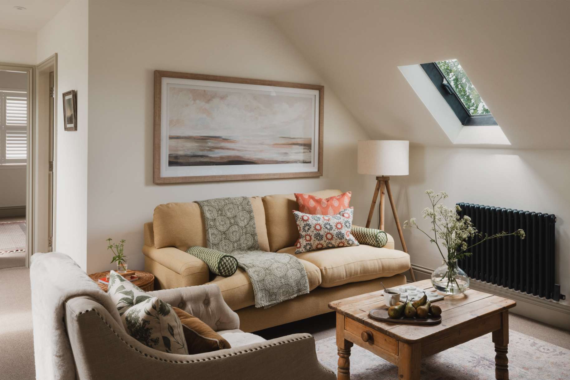 A mustard sofa with patterned cushions sits beneath a skylight, an armchair and wooden coffee table with a vase of flowers in the foreground.