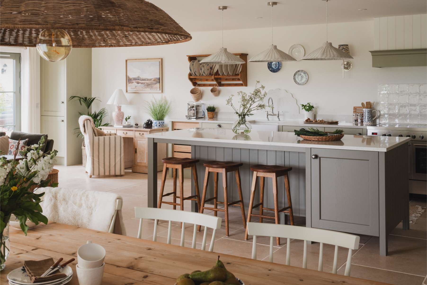 A kitchen island with wooden stools stands before a sink and range, hanging pendant lights and a woven light shade above the dining table in the foreground.