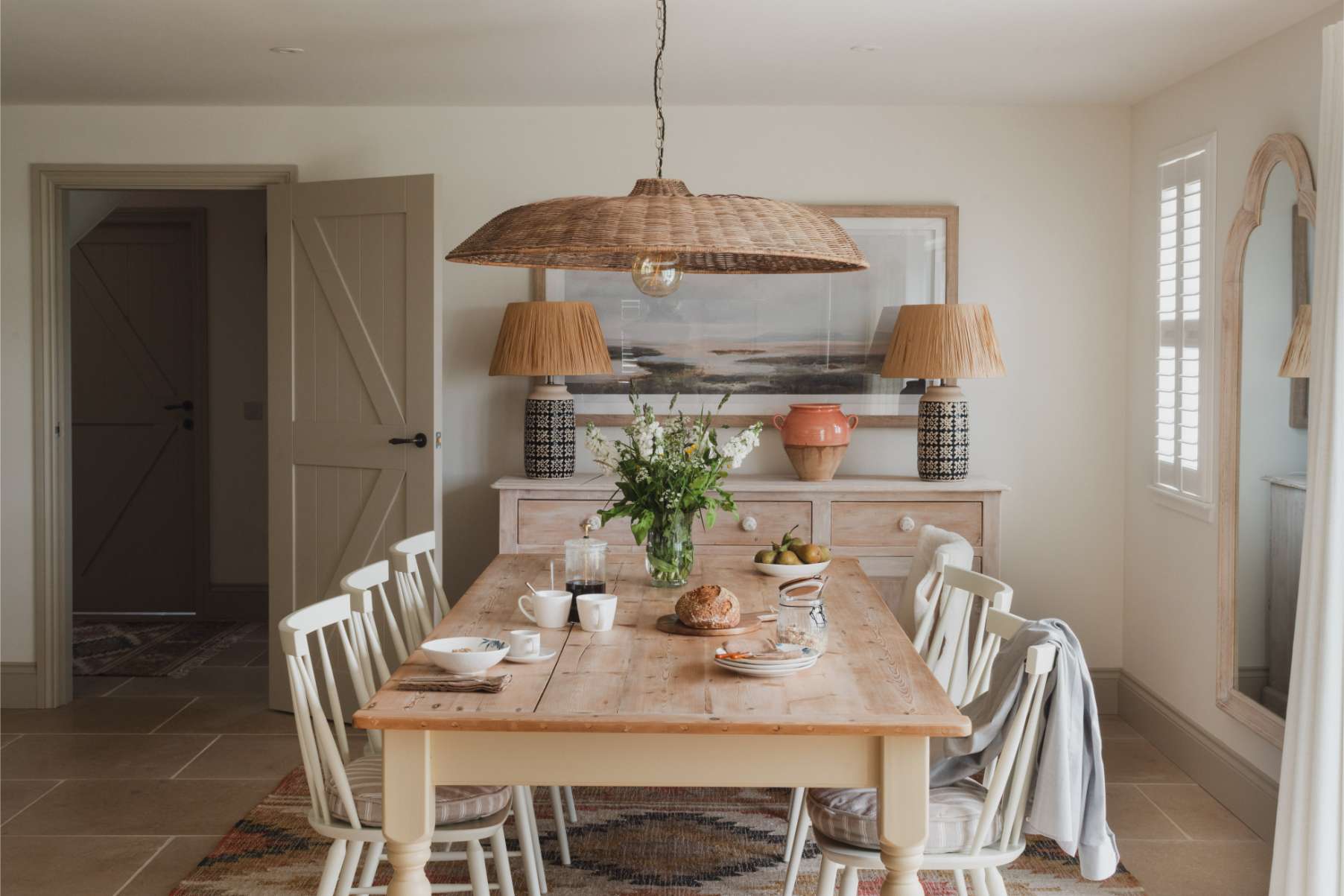 A dining table set with fruit, bread and flowers stands before a wicker pendant light and a sideboard with lamps and a framed print.