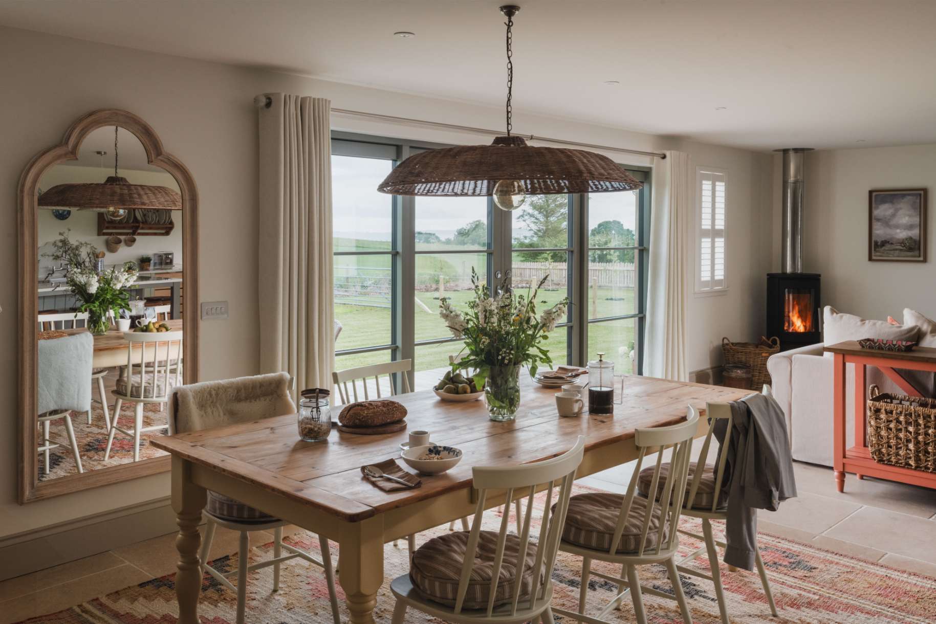 A wooden dining table set with flowers stands before glazed doors looking onto a field, a wood burning stove and mirror to the right.