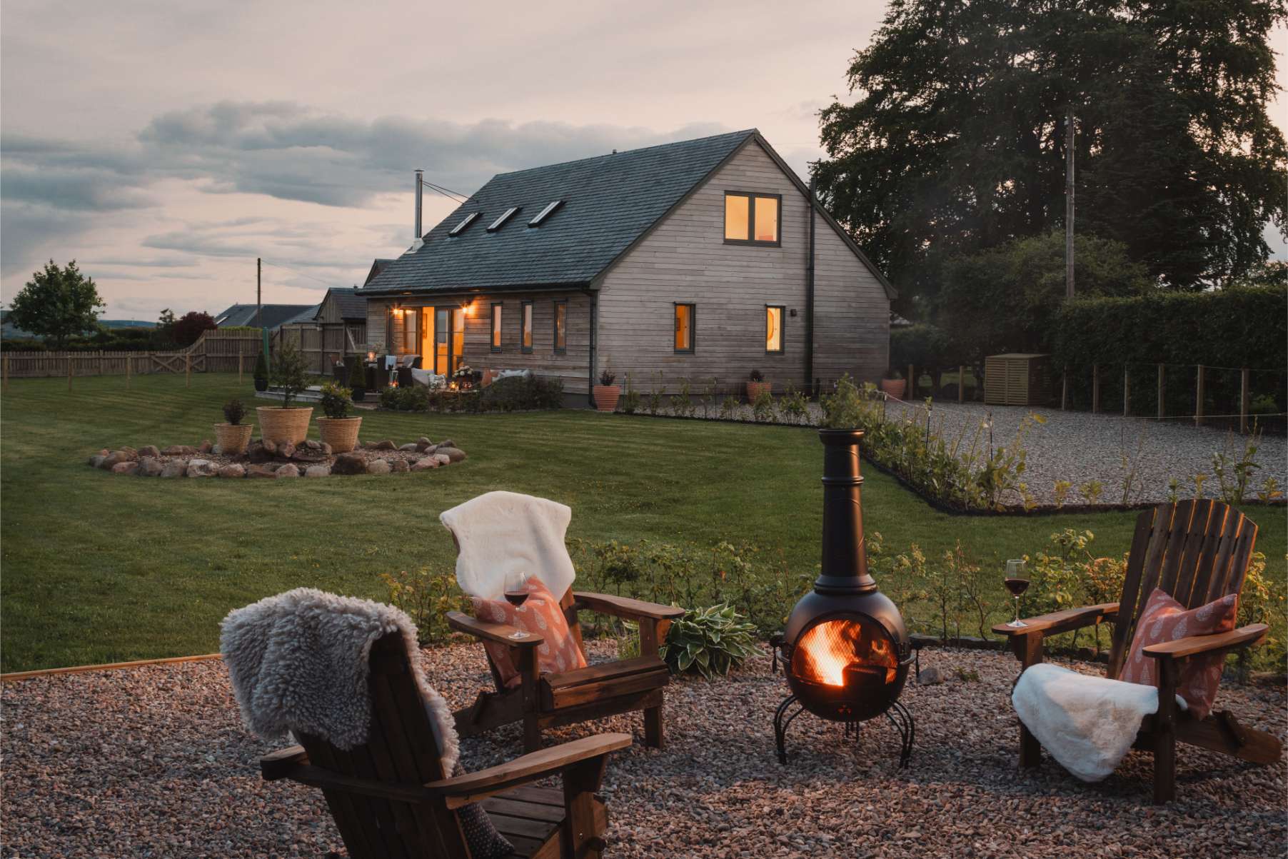 Four wooden adirondack chairs draped with throws circle a lit fire bowl on a gravel patio, a timber house glowing at dusk behind.