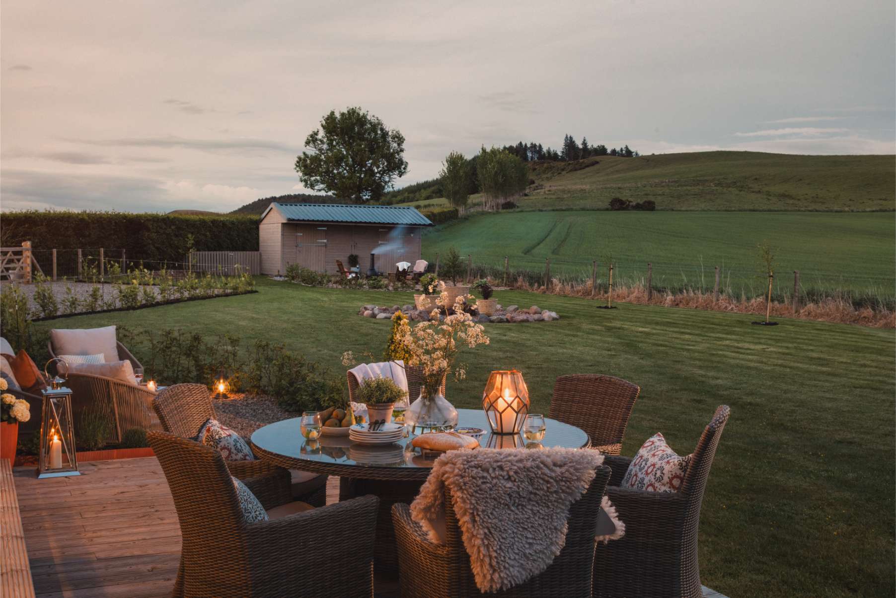 A wicker dining table set with candles and flowers stands on a deck at dusk, rolling fields stretching towards a distant barn.
