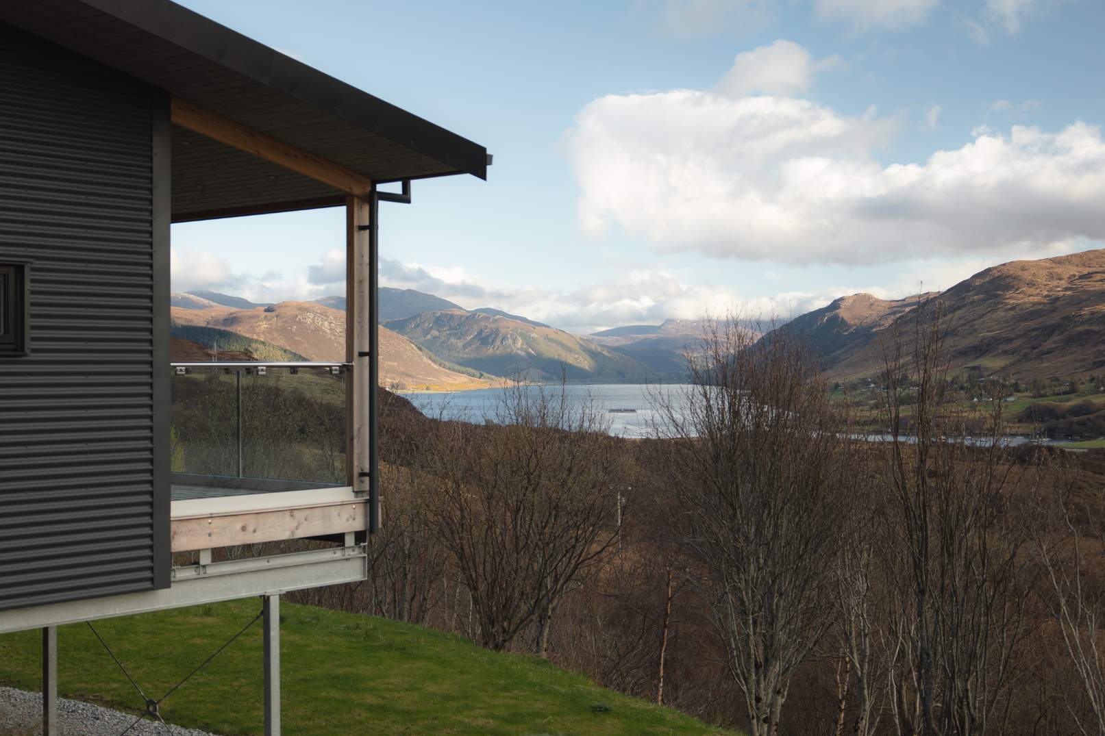 The timber-clad house's cantilevered corner overlooks a loch framed by hills, bare trees in the foreground.